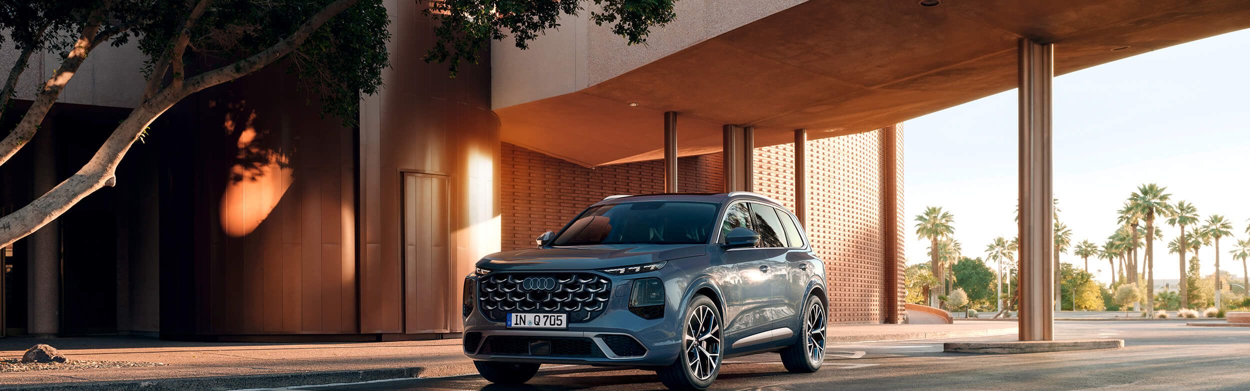 Sodoben športni terenec Audi Q7 stoji na cesti pred elegantno, sodobno stavbo z velikim nadstreškom. Sončna svetloba ustvarja dolge sence avtomobila na asfaltu. V ozadju so palme in modro nebo, kar daje prizoru občutek toplote in prestiža.