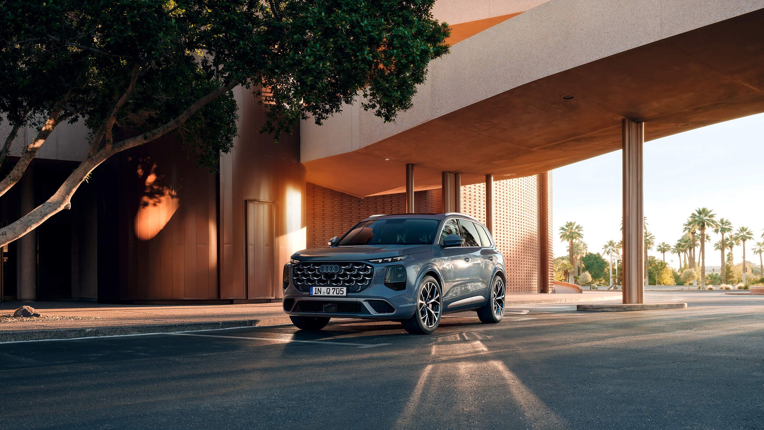 Sodoben športni terenec Audi Q7 stoji na cesti pred elegantno, sodobno stavbo z velikim nadstreškom. Sončna svetloba ustvarja dolge sence avtomobila na asfaltu. V ozadju so palme in modro nebo, kar daje prizoru občutek toplote in prestiža.