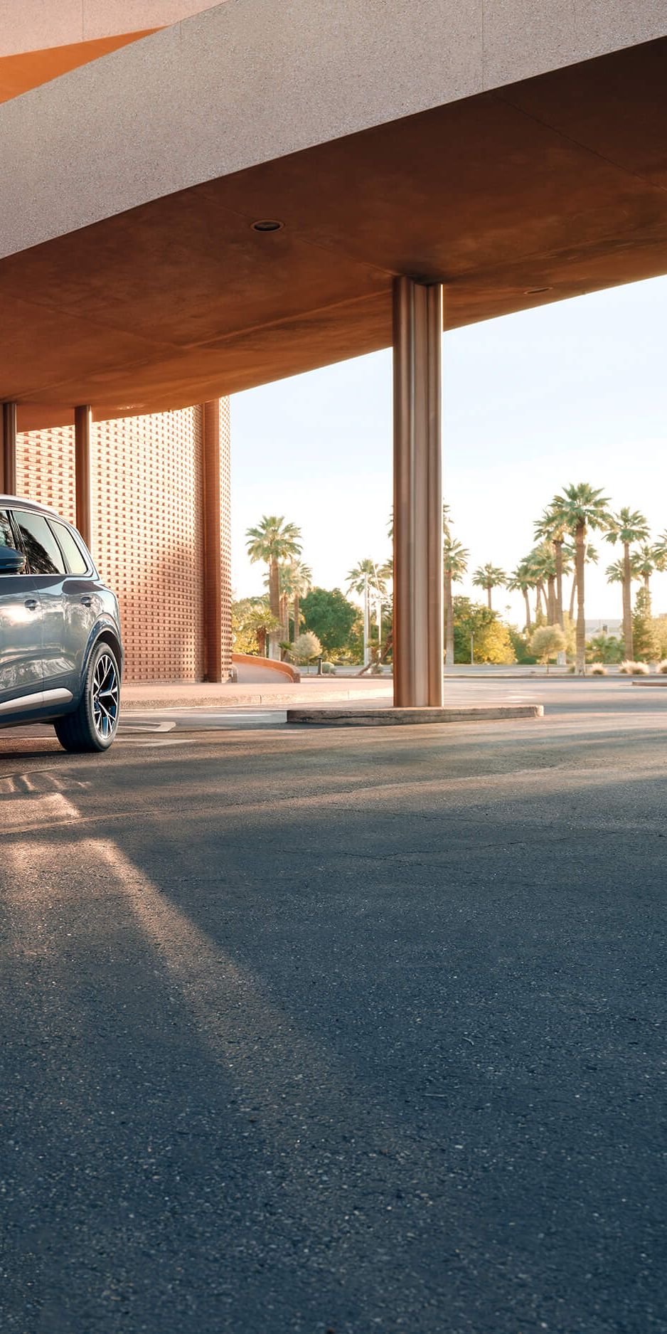 Sodoben športni terenec Audi Q7 stoji na cesti pred elegantno, sodobno stavbo z velikim nadstreškom. Sončna svetloba ustvarja dolge sence avtomobila na asfaltu. V ozadju so palme in modro nebo, kar daje prizoru občutek toplote in prestiža.