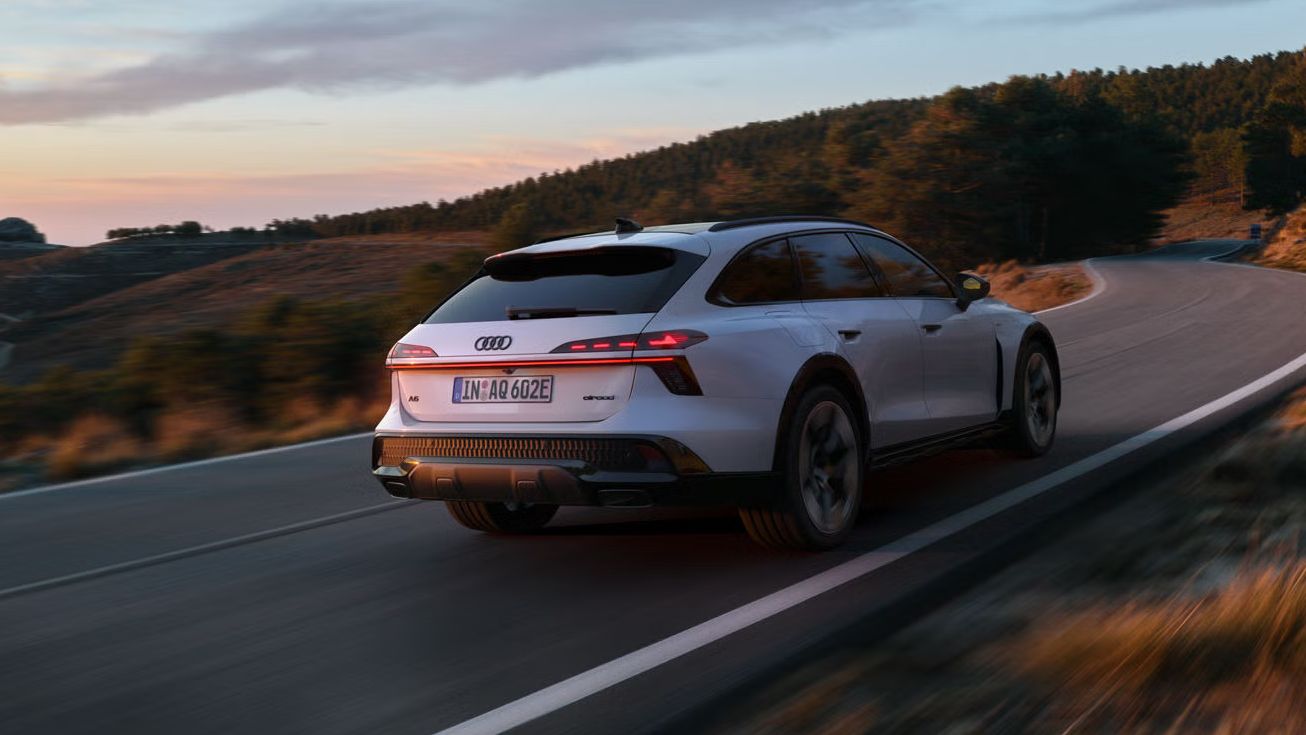 Audi A6 allroad e‑hybrid, prikazan diagonalno od zadaj pri polni hitrosti na gorski cesti; v ozadju se vidi del gozda.
