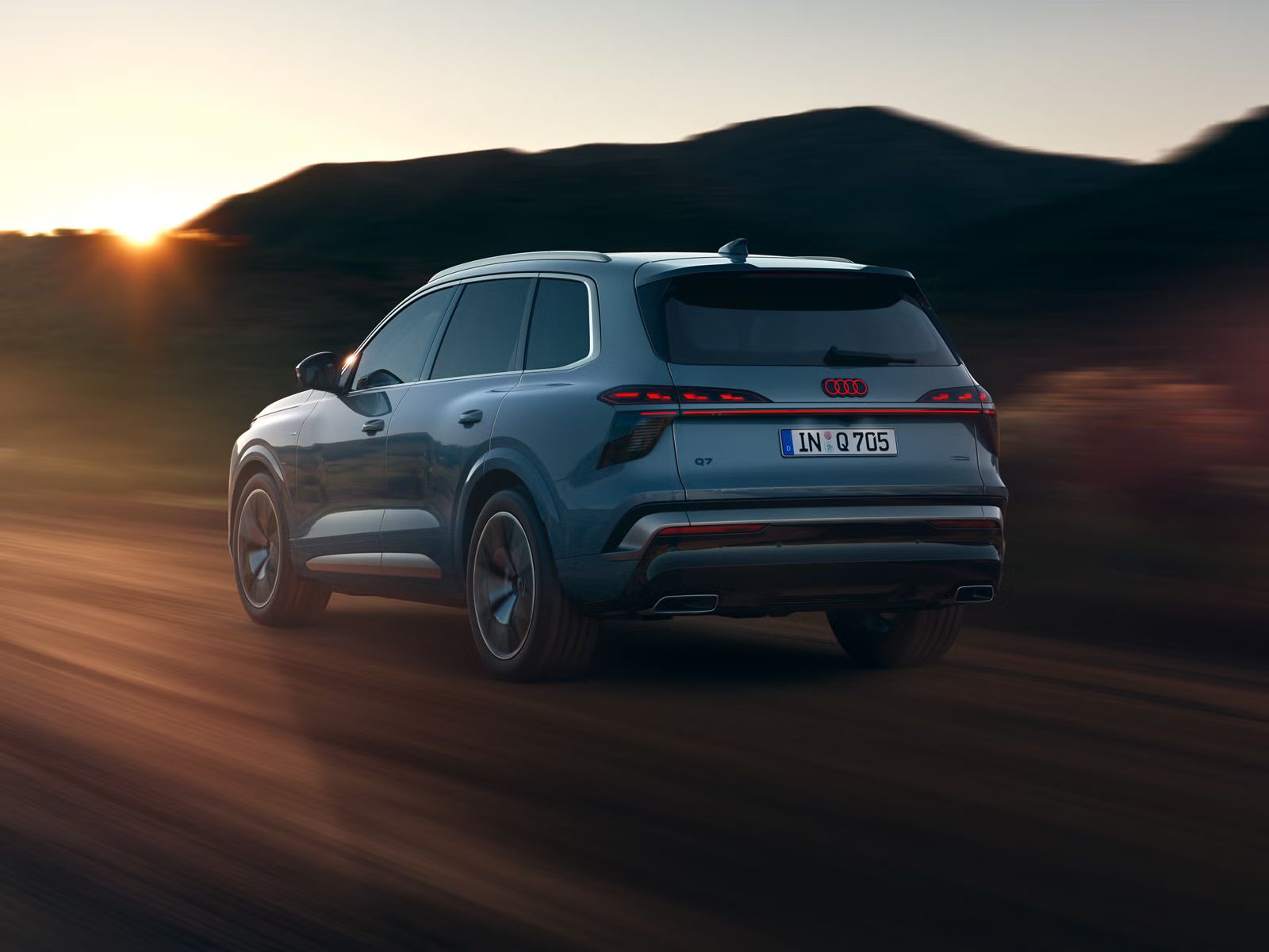 Der Audi Q7 SUV schräg von hinten in voller Fahrt auf einer Landstraße, im Hintergrund Gebirge. Die am Horizont untergehende Sonne beleuchtet die Szenerie.  