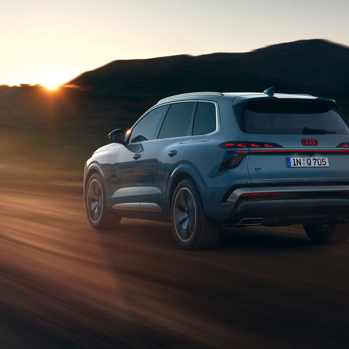 Audi Q7 SUV diagonalno od zadaj med vožnjo po podeželski cesti, v ozadju gore. Sonce, ki zahaja na obzorju, osvetljuje pokrajino.