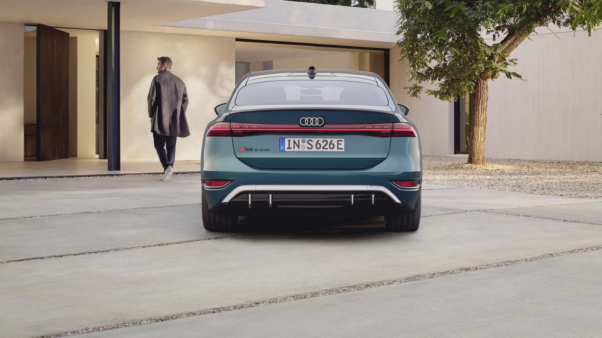 Audi S6 Sportback e-tron, fotografiran od zadaj, parkiran pred sodobno hišo z ravnimi linijami in svetlimi fasadami. V ozadju moški hodi proti vhodu v hišo, medtem ko zadnji del avtomobila poudarjajo neprekinjena LED svetlobna linija, športni difuzor in oznaka S6 e-tron.