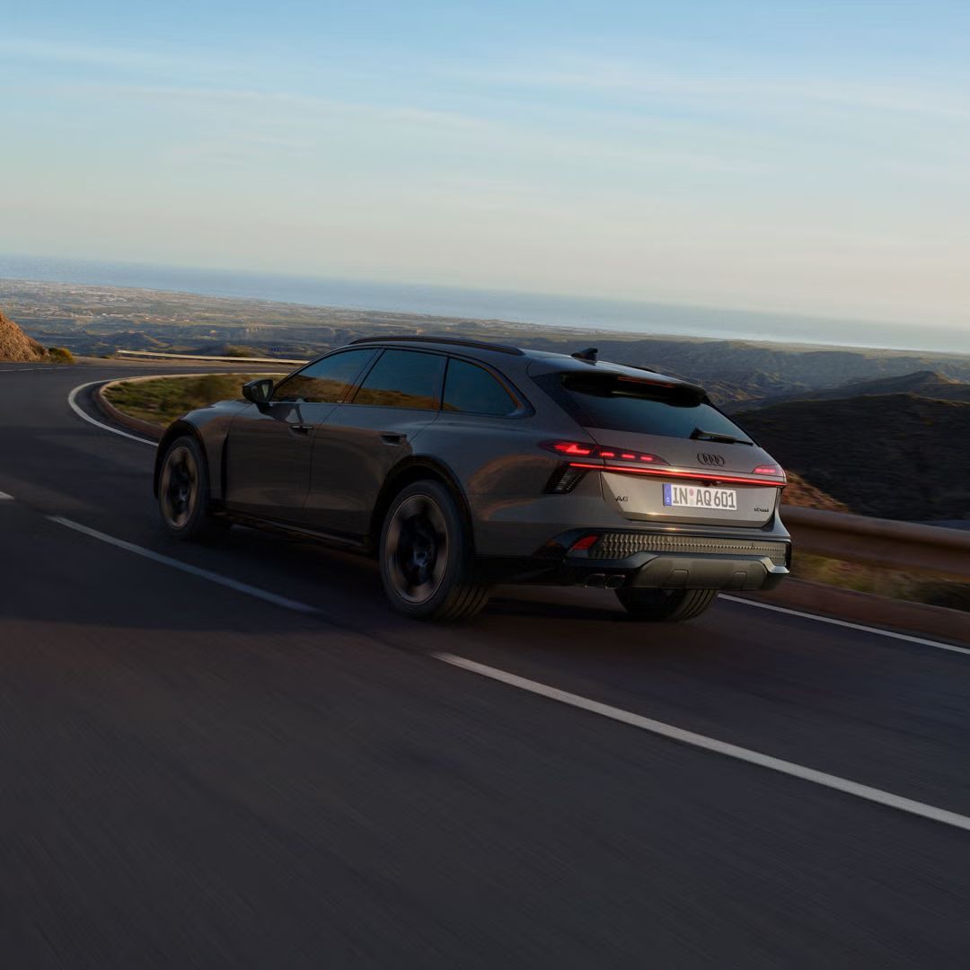 Audi A6 allroad diagonalno od zadaj vozi skozi dolg ovinek po podeželski cesti.