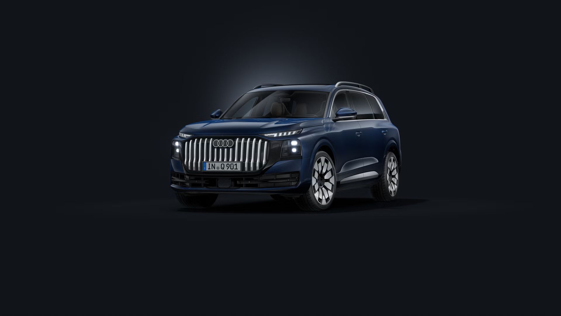 Temno moder Audi Q9 SUV, fotografiran iz sprednjega bočnega kota na temnem studijskem ozadju, z izrazito enodelno masko hladilnika, markantnim oblikovanjem in velikimi aluminijastimi platišči.