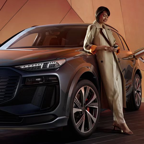 Vozilo Audi SQ6 je priključeno na električni polnilnik, na vozilo je naslonjena ženska, ki brska po telefonu.