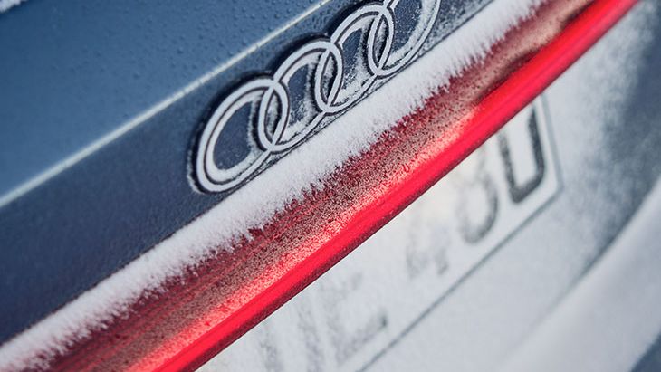 Na zadnjem delu avtomobila Audi S5, pokritem z ivjem, je viden znak blagovne znamke Audi. Nad rdečo črto zadnje luči je logotip s štirimi povezanimi krogi. Spodaj je registrska tablica, delno prekrita s snegom.