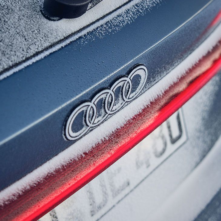 Na zadnjem delu avtomobila Audi S5, pokritem z ivjem, je viden znak blagovne znamke Audi. Nad rdečo črto zadnje luči je logotip s štirimi povezanimi krogi. Spodaj je registrska tablica, delno prekrita s snegom.