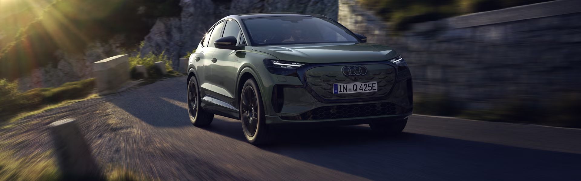 Audi Q4 Sportback e-tron med vožnjo po gorski cesti. Sliko z leve zgornje strani osvetljujejo sončni žarki, vozilo meče senco predse.