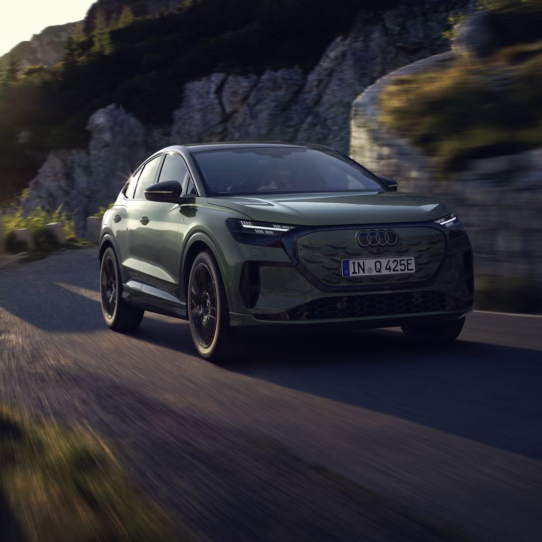 Audi Q4 Sportback e-tron med vožnjo po gorski cesti. Sliko z leve zgornje strani osvetljujejo sončni žarki, vozilo meče senco predse.