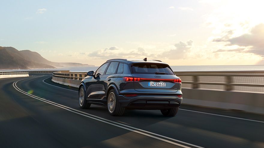 Temno moder Audi Q6 e-tron vozi po zaviti obalni cesti v sončnem vremenu. Avto je fotografiran od zadaj, na desni strani je ograja mostu. V ozadju so morje, hribi in delno oblačno nebo z mehko popoldansko svetlobo.