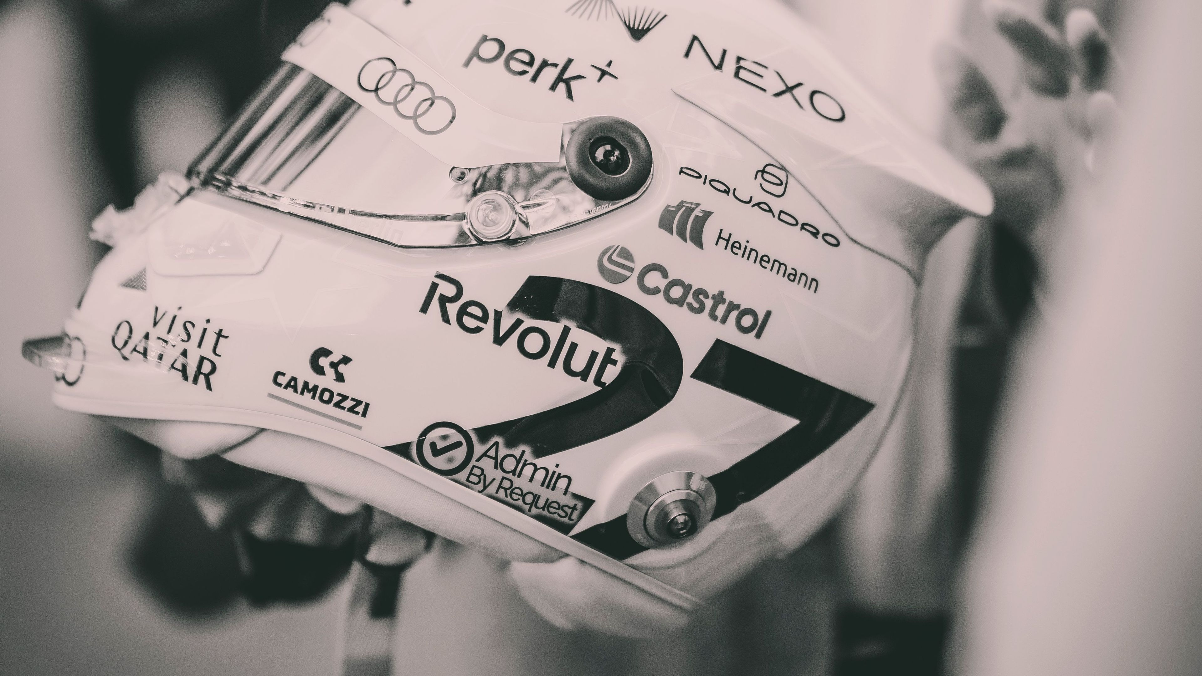 Oseba v elegantni obleki drži belo dirkaško čelado, prekrito z logotipi različnih sponzorjev, kot so Revolut, Audi in Castrol. Fotografija je v črno-beli oziroma sepia tehniki, kar daje prizoru eleganten, nekoliko nostalgičen videz. Ozadje je mehko zamegljeno, poudarek je na čeladi v ospredju.