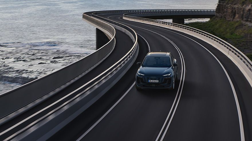 Temno moder avtomobil Audi vozi po široki, dvopasovni cesti, ki se dramatično vije ob obali z razgledom na morje. Cesta je dvignjena nad skalnato obalo, z visokimi varnostnimi ograjami na obeh straneh. Prizor deluje dinamično in turistično.