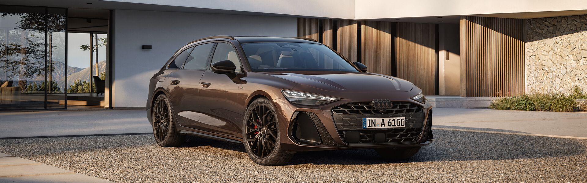 Rjavo Audi A6 vozilo stoji na dvorišču pred modernim stanovanjem. Avto ima črno platišče in športni izgled. Ozadje sestavljajo bela in lesena fasada hiše.