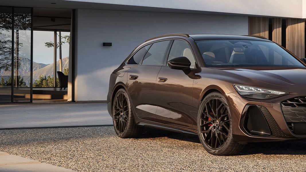 Rjavo Audi A6 vozilo stoji na dvorišču pred modernim stanovanjem. Avto ima črno platišče in športni izgled. Ozadje sestavljajo bela in lesena fasada hiše.