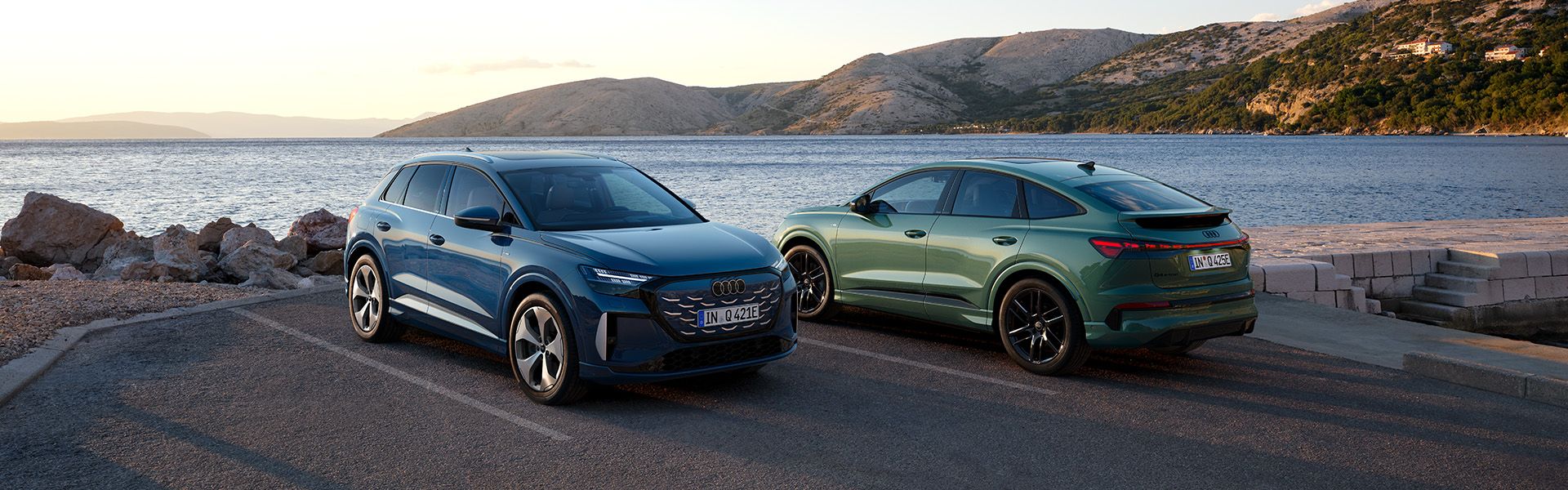 Audi Q4 SUV e-tron quattro in Audi Q4 Sportback e-tron stojita parkirani ob morju, eno v modri in drugo v zeleni barvi. V ozadju so mirno morje, skalnata obala in hribovit teren pod mehko svetlobo sončnega zahoda. Scena poudarja eleganten dizajn avtomobilov v naravnem, sproščenem okolju.