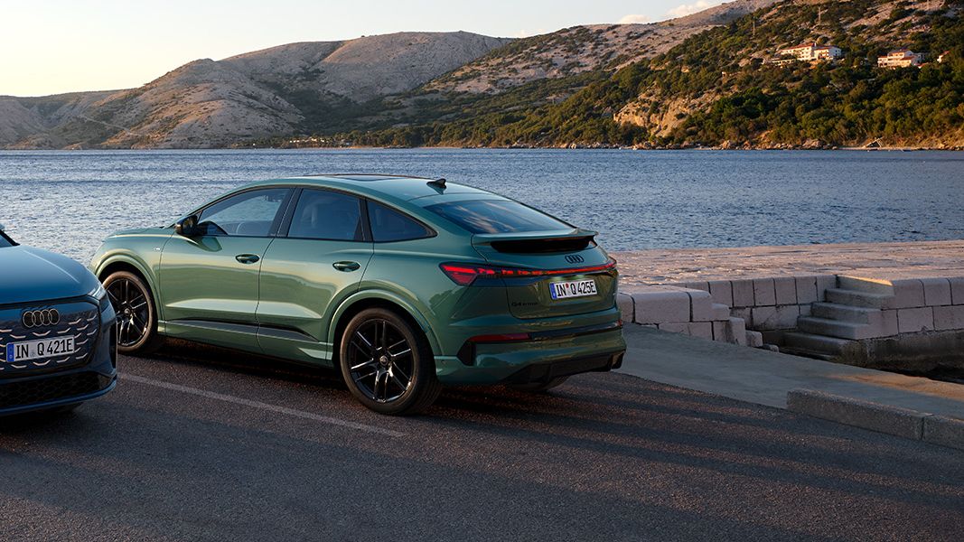 Audi Q4 SUV e-tron quattro in Audi Q4 Sportback e-tron stojita parkirani ob morju, eno v modri in drugo v zeleni barvi. V ozadju so mirno morje, skalnata obala in hribovit teren pod mehko svetlobo sončnega zahoda. Scena poudarja eleganten dizajn avtomobilov v naravnem, sproščenem okolju.