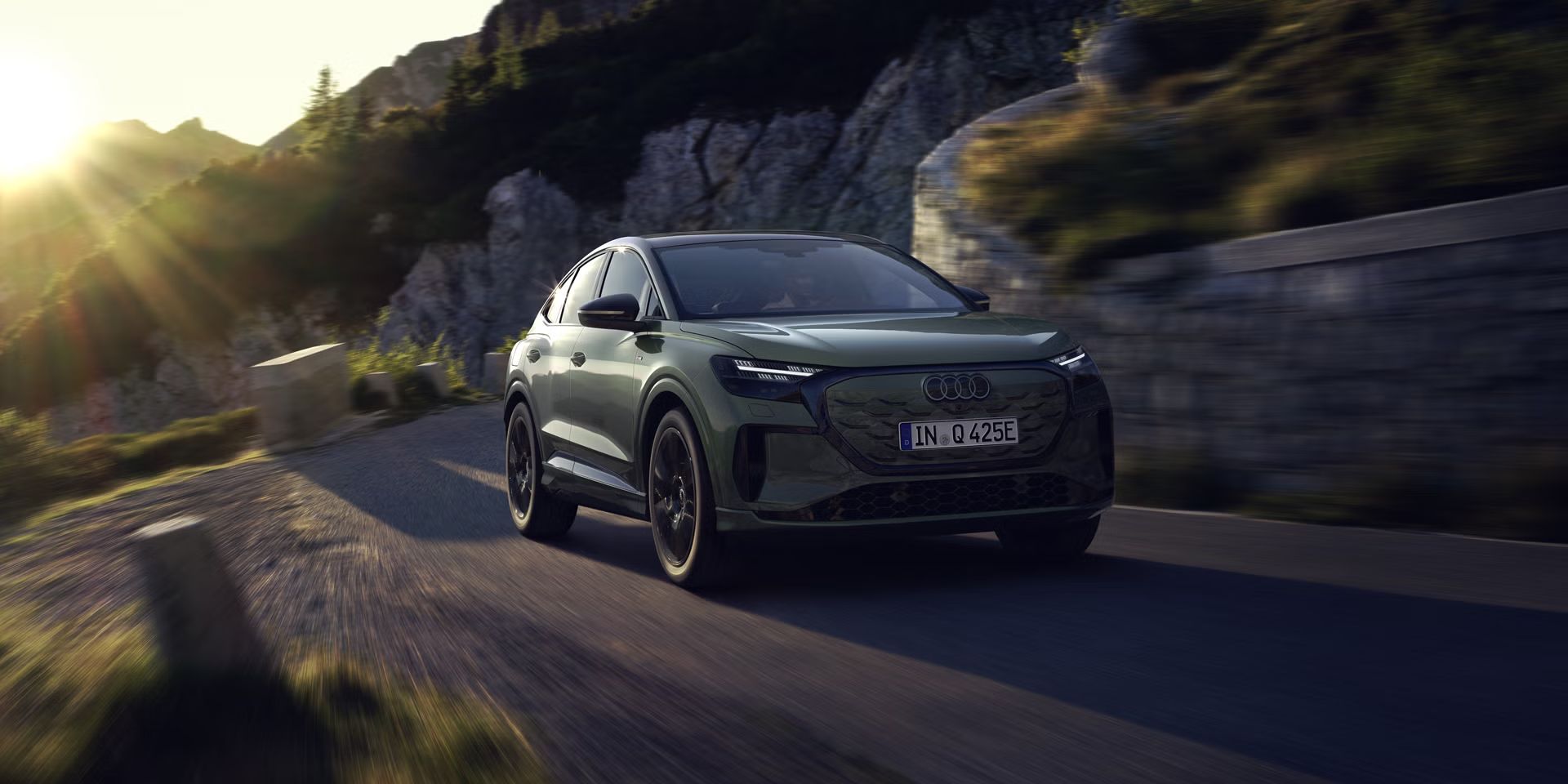 Audi Q4 Sportback e-tron med vožnjo po gorski cesti. Sliko z leve zgornje strani osvetljujejo sončni žarki, vozilo meče senco predse.