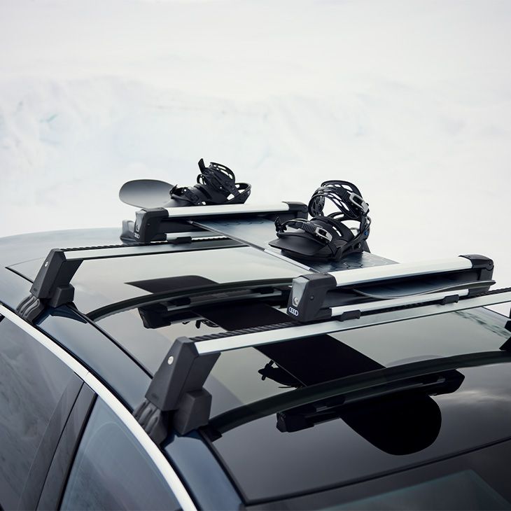 Strešni nosilec z nosilci za snežno desko, nameščen na strehi vozila Audi.  Snežna deska je varno pritrjena na prečnih nosilcih s pritrdilnimi elementi.  Posnetek prikazuje transport športne opreme v zimskem, zasneženem okolju.