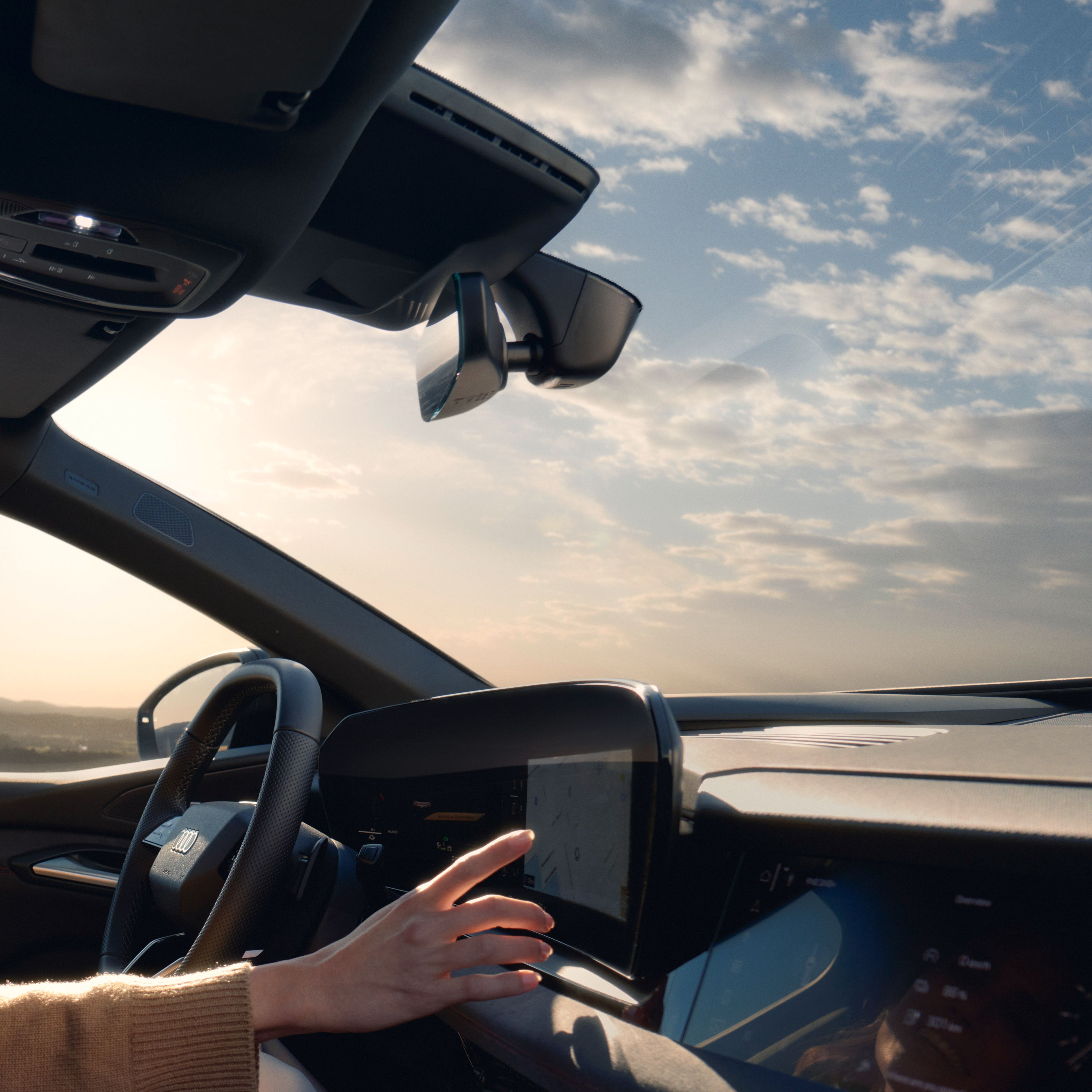 Oseba v avtomobilu dotika osrednji zaslon na armaturni plošči, medtem ko je viden volan in del vetrobranskega stekla. Zunaj skozi okno je čudovito jasno nebo z nekaj oblački.