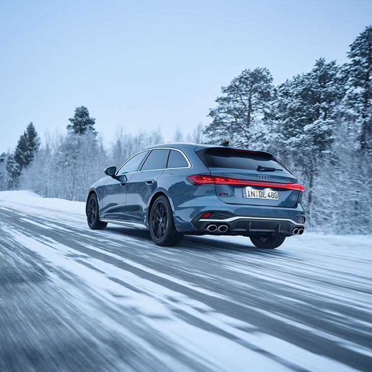 Modra Audi S5 limuzina vozi po zasneženi cesti skozi zimsko pokrajino. Cesta je obdana s sneženimi drevesi. Vozilo ima nemške registrske tablice.