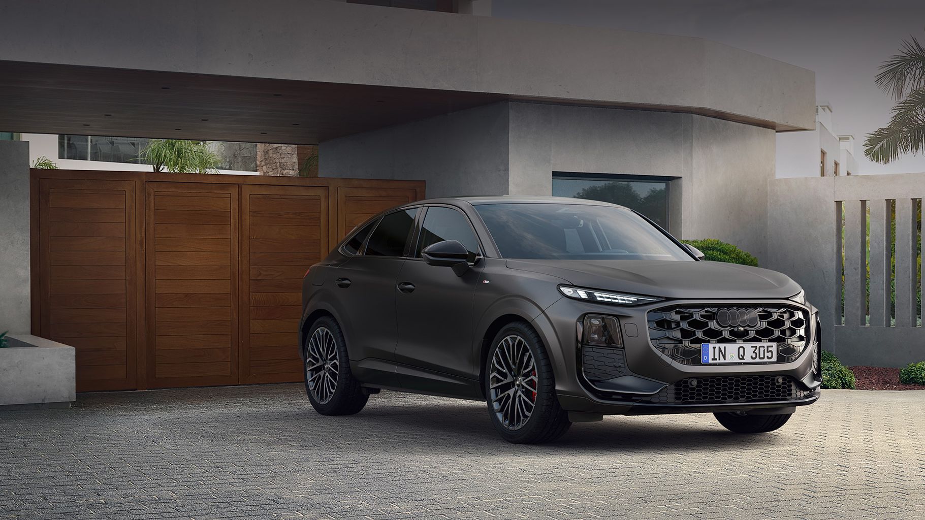 Temno siv športni Audi Q3 Sportback parkiran pred hišo z lesenimi garažnimi vrati. Avtomobil ima eleganten dizajn in velika platišča. V ozadju so vidne palme in okenska stekla hiše.