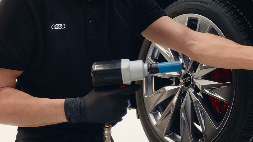 Mehanik v delavnici popravlja pnevmatiko na vozilu Audi. Uporablja električno orodje za privijanje vijakov na platišču avtomobila. Mehanik ima oblečeno črno majico z logotipom Audi.