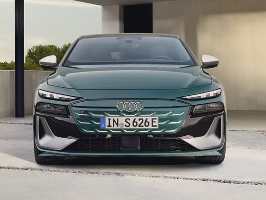 Audi S6 Sportback e-tron, fotografiran od spredaj, v temno zeleni barvi, parkiran pred sodobno minimalistično hišo. Avtomobil ima široko in nizko držo, izrazito sprednjo masko z osvetljenimi detajli ter ostre LED žaromete, ki poudarjajo njegov športen in električen značaj.