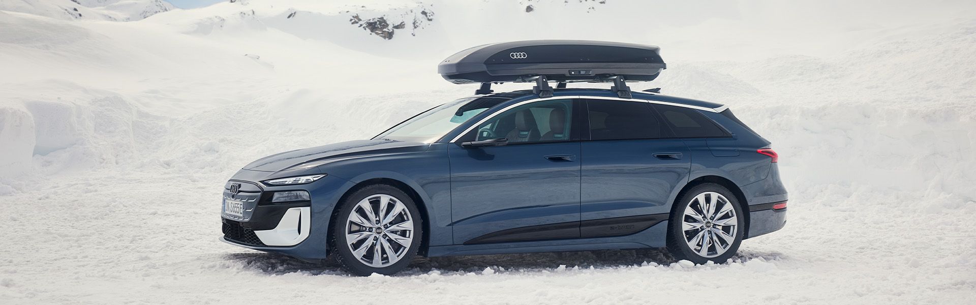 Audi A6 Avant v temno modrem odtenku, prikazan v bočnem pogledu, parkiran v zasneženem gorskem okolju.  Na strehi je nameščen strešni kovček, vidna so tudi platišča in zaščitni spodnji elementi karoserije.  Prizor jasno prikazuje vozilo v zimskih razmerah ter njegovo postavitev v odprtem, naravnem prostoru.