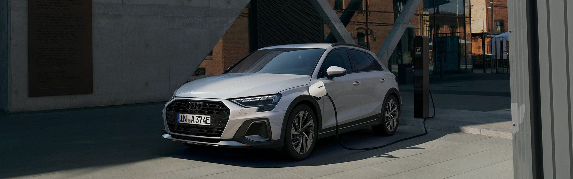 Audi A3 allstreet e-hybrid je priključen na polnilno postajo in stoji na sodobnem parkirišču. Polnilni kabel je povezan z levim bokom vozila, ozadje pa zaznamujejo moderna steklena in opečnata poslopja.