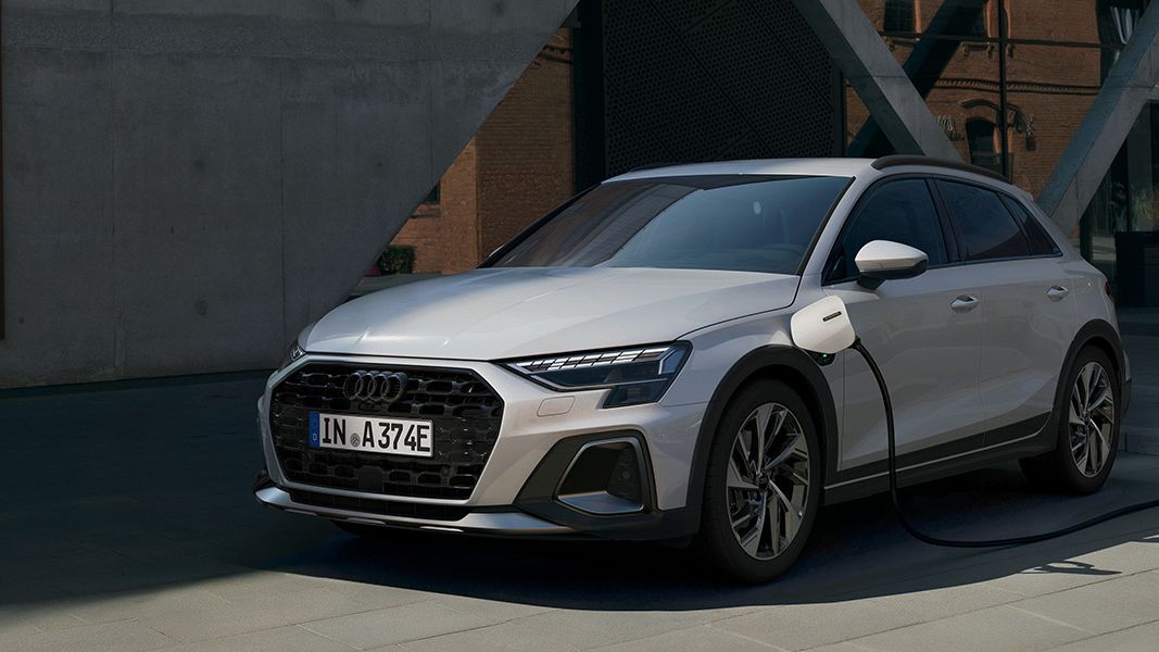 Audi A3 allstreet e-hybrid je priključen na polnilno postajo in stoji na sodobnem parkirišču. Polnilni kabel je povezan z levim bokom vozila, ozadje pa zaznamujejo moderna steklena in opečnata poslopja.