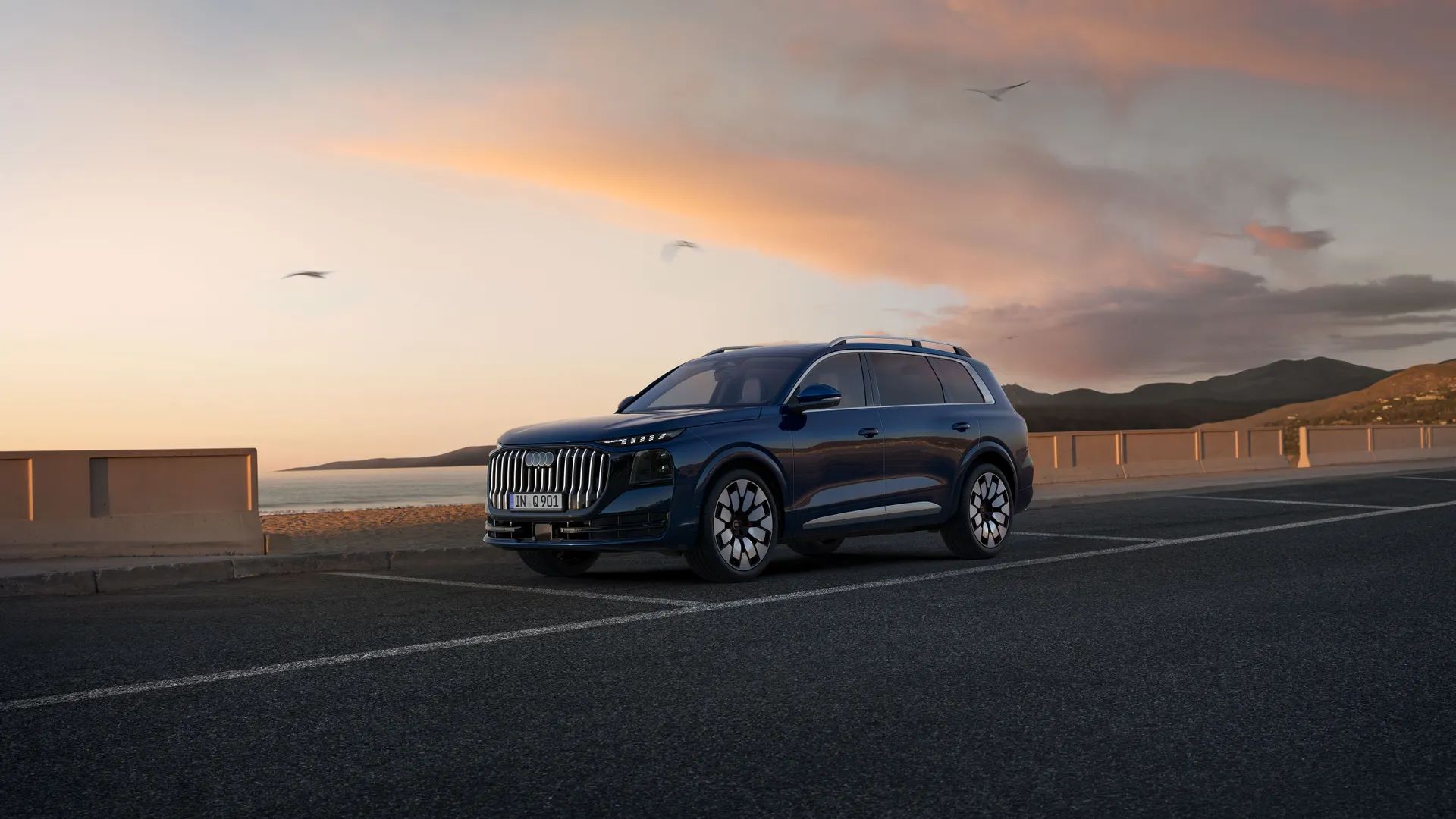 Temno moder Audi Q9 je parkiran ob obmorski promenadi, v ozadju pa je mirno morje in pastelno obarvano nebo ob sončnem zahodu.  