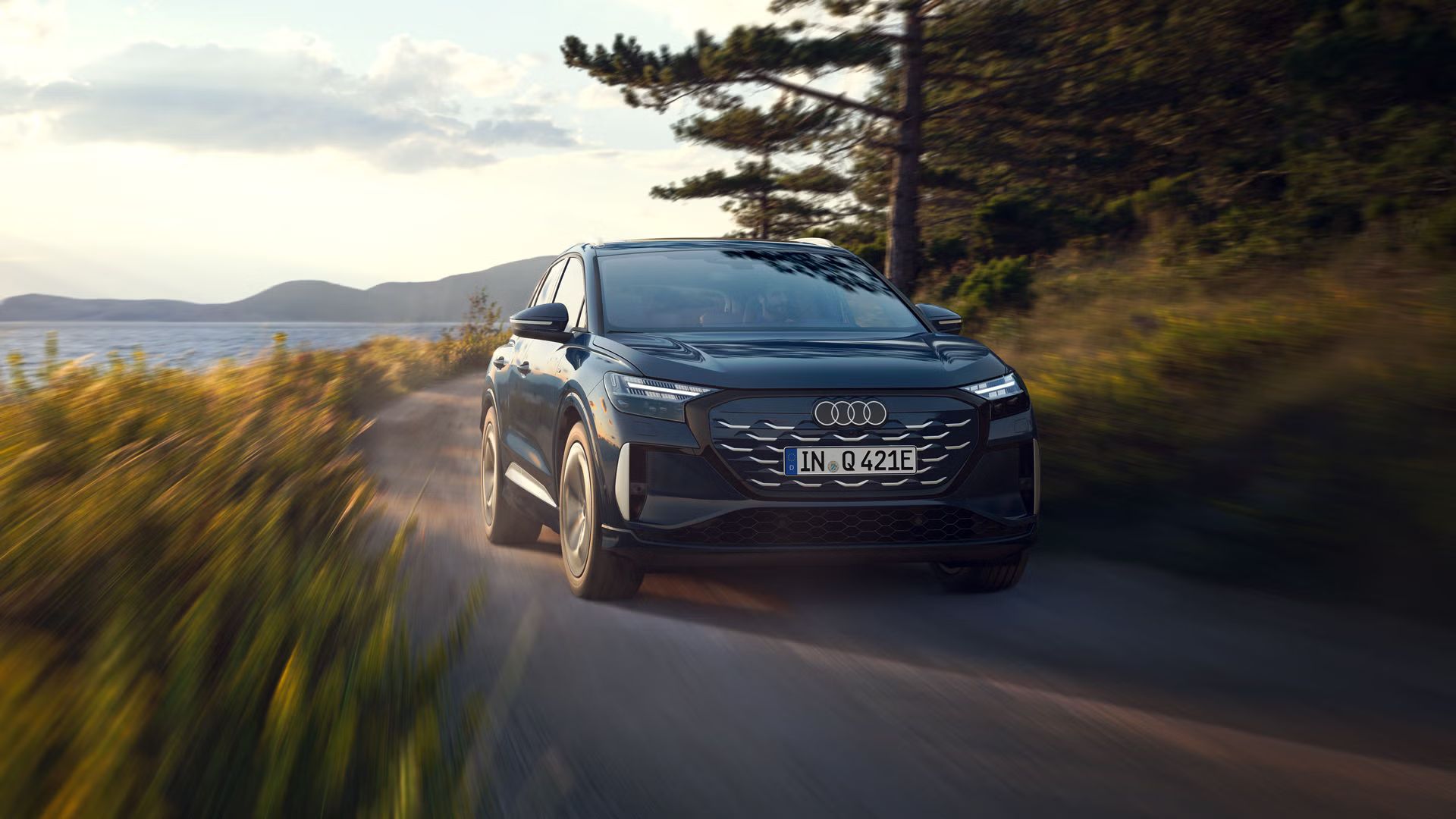 Stranski sprednji del vozila Audi Q4 SUV e-tron pri vožnji po regionalni cesti ob gozdnem jezeru, v ozadju gorska pokrajina.