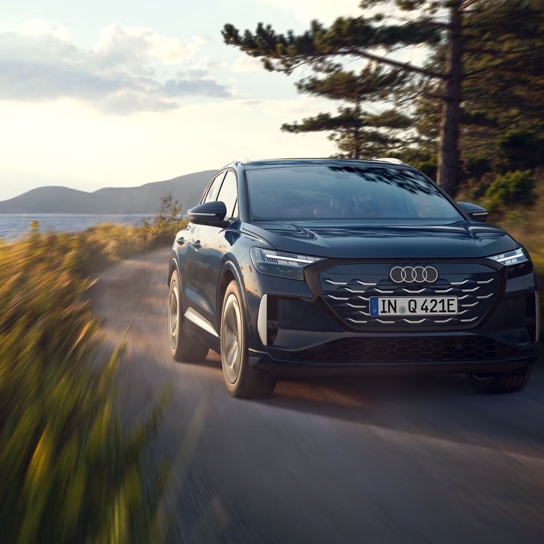 Stranski sprednji del vozila Audi Q4 SUV e-tron pri vožnji po regionalni cesti ob gozdnem jezeru, v ozadju gorska pokrajina.