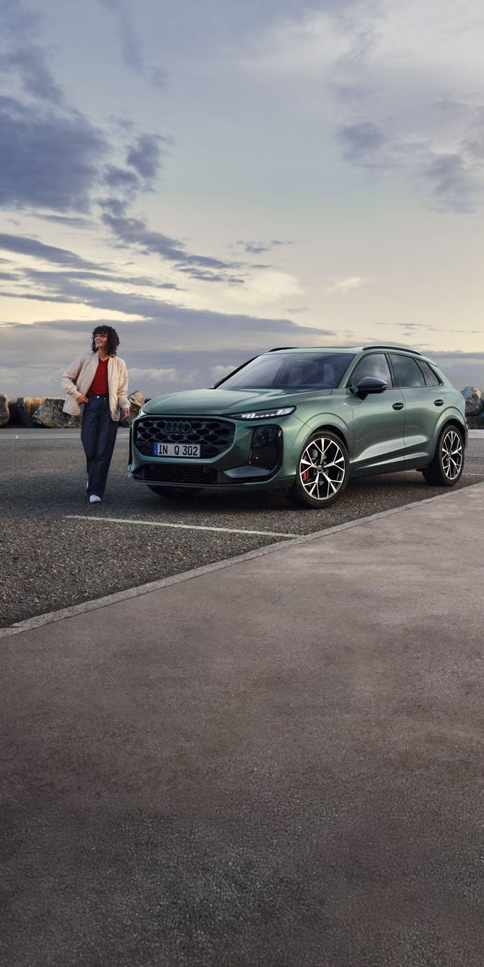 Seitliche, vordere Ansicht auf den geparkten Q3 SUV an einer Küstenstraße, daneben eine junge Frau. 