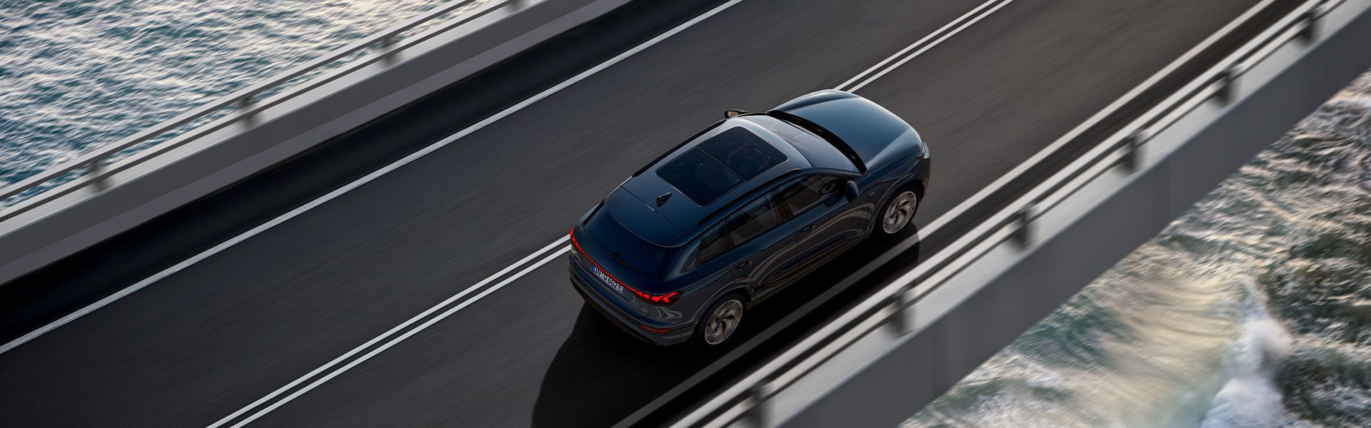 Temno moder Audi Q6 e-tron vozi po viaduktu nad morjem, posnet od zgoraj. Desno spodaj se vidijo valovi, ki butajo ob skale, levo pa mirnejša morska gladina. Cesta je dvopasovna z belimi črtami in kovinsko ograjo.