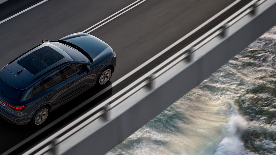 Temno moder Audi Q6 e-tron vozi po viaduktu nad morjem, posnet od zgoraj. Desno spodaj se vidijo valovi, ki butajo ob skale, levo pa mirnejša morska gladina. Cesta je dvopasovna z belimi črtami in kovinsko ograjo.