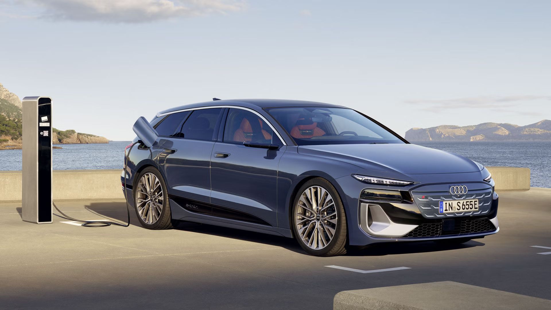 Električni Audi je parkiran ob polnilni postaji in priključen na polnjenje. Vozilo je prikazano s strani, ob obalni cesti z morjem in hribi v ozadju.