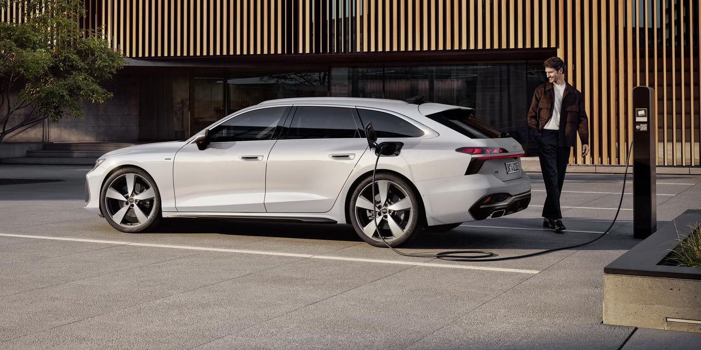 Audi A6 Avant e-hybrid v beli barvi, parkiran ob polnilni postaji pred sodobno stavbo z leseno fasado.  Vozilo je priključeno na polnjenje, polnilni kabel je vstavljen v zadnji bočni del avtomobila.  Ob vozilu stoji moški, prizor pa jasno prikazuje vsakdanjo uporabo elektrificiranega pogona v urbanem okolju.