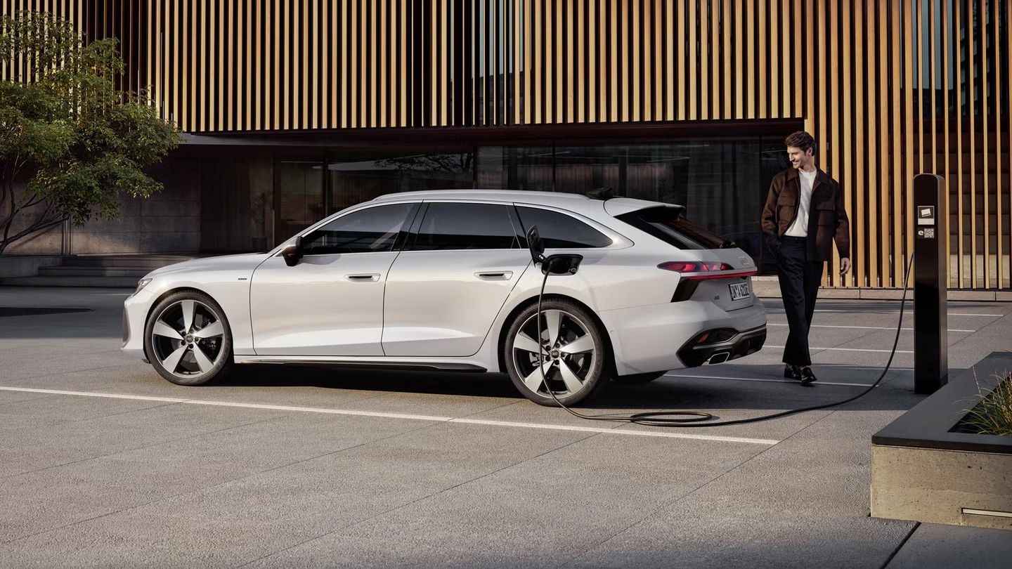 Audi A6 Avant e-hybrid v beli barvi, parkiran ob polnilni postaji pred sodobno stavbo z leseno fasado.  Vozilo je priključeno na polnjenje, polnilni kabel je vstavljen v zadnji bočni del avtomobila.  Ob vozilu stoji moški, prizor pa jasno prikazuje vsakdanjo uporabo elektrificiranega pogona v urbanem okolju.