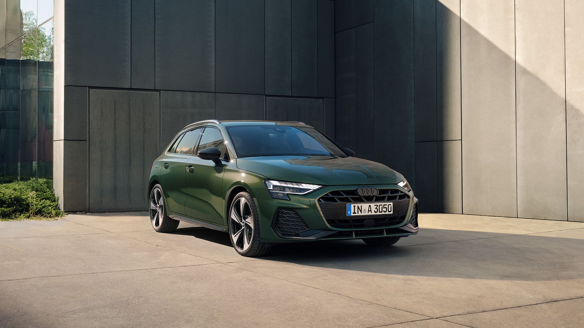 Audi A3 Sportback, parkiran pred sodobno arhitekturno fasado.  Vozilo je temno zelene barve, s poudarjenimi LED žarometi in aerodinamično oblikovanim sprednjim delom.  Okolje je urbano in umirjeno, z igro svetlobe in senc, ki poudarja linije vozila.