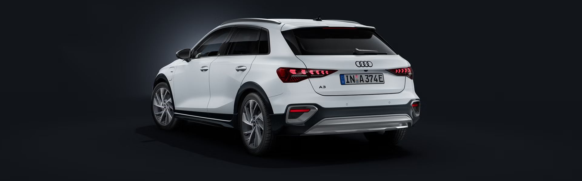 Audi A3 allstreet TFSI e v beli barvi, prikazan iz zadnjega bočnega kota na temnem ozadju. Izrazite LED-zadnje luči, zaščitni elementi odbijača, strešna nosilca in priključnohibridni pogon poudarjajo sodoben dizajn, učinkovitost in vsestranskost vozila.