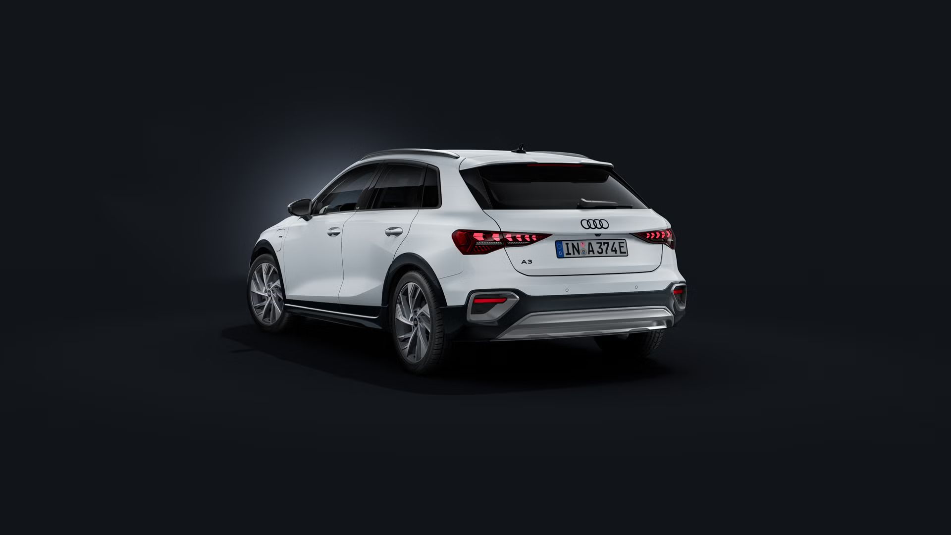 Audi A3 allstreet TFSI e v beli barvi, prikazan iz zadnjega bočnega kota na temnem ozadju. Izrazite LED-zadnje luči, zaščitni elementi odbijača, strešna nosilca in priključnohibridni pogon poudarjajo sodoben dizajn, učinkovitost in vsestranskost vozila.